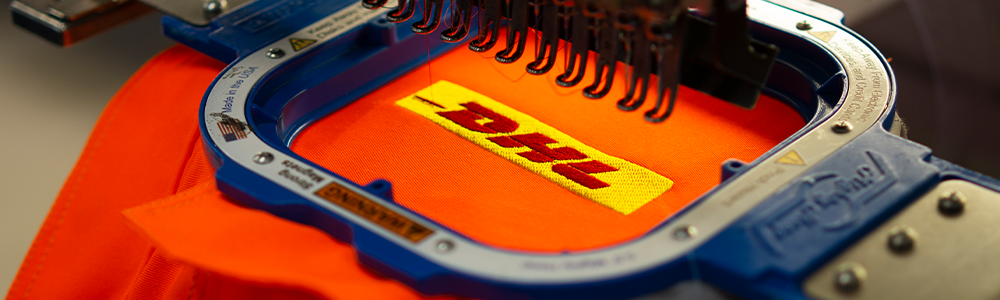 Borduur logo Borduur machine met een lichte oranje stof ingeklemd in een ring met een geel logo.